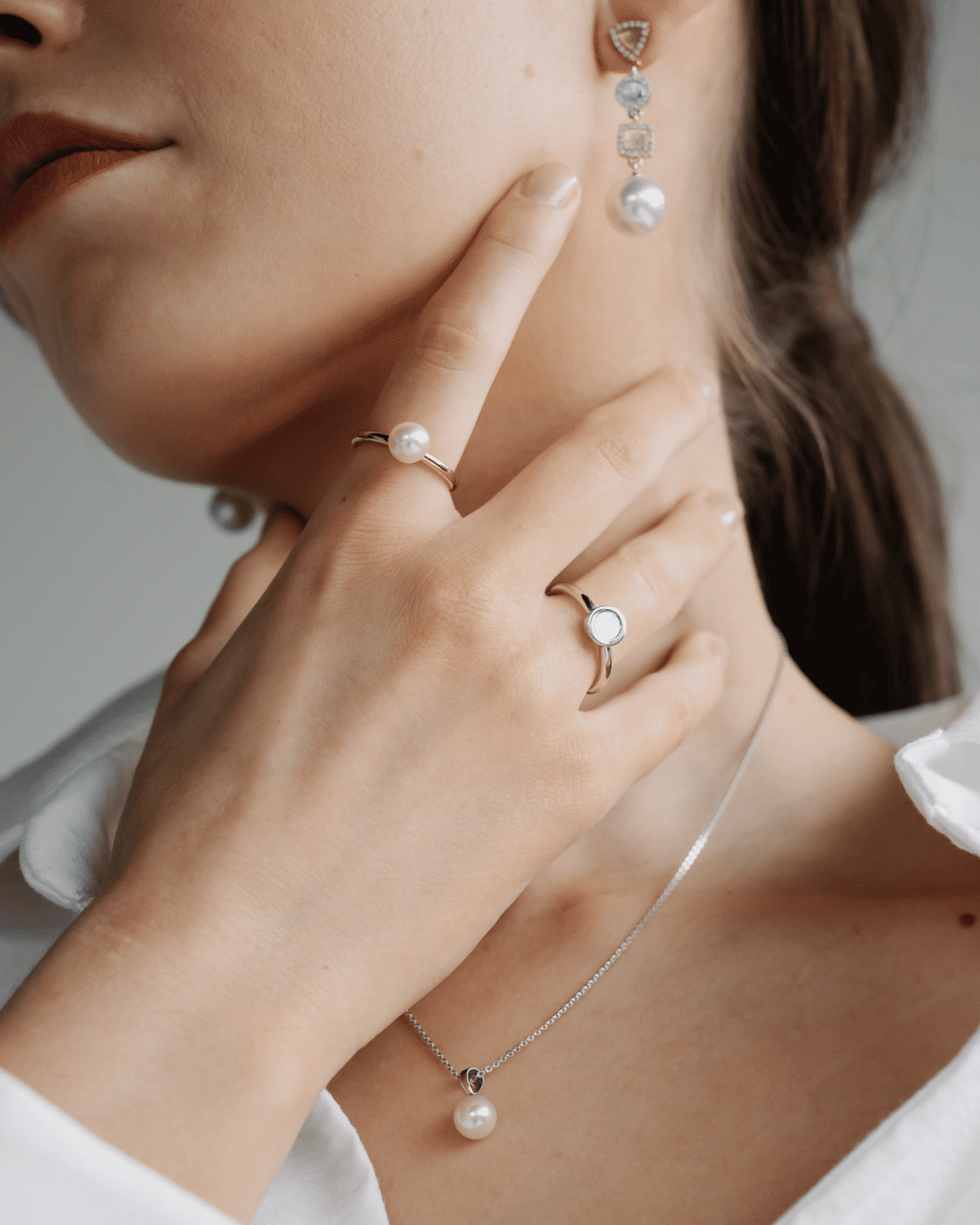 A woman wears her stunning pearl and mother of pearl rings, a classic pearl pendant on a dainty chain, and a pair of statement pearl earrings featuring geometric shapes and sparkling, white diamonds.