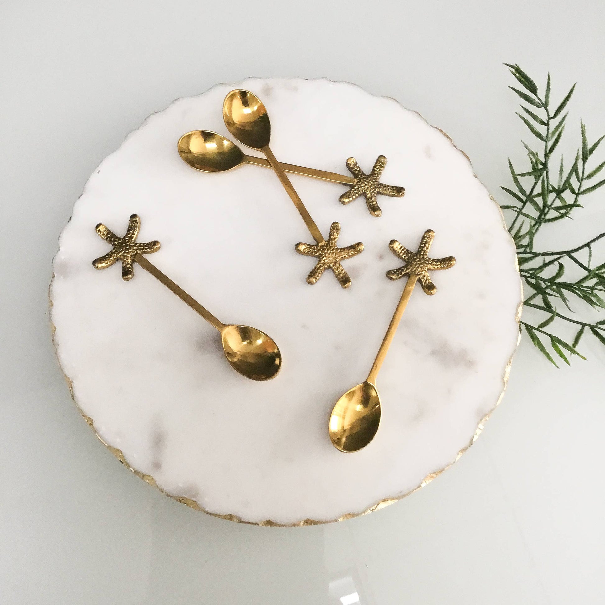 Set of gold spoons shaped like starfish on a white plate with a green leafy branch in the background.