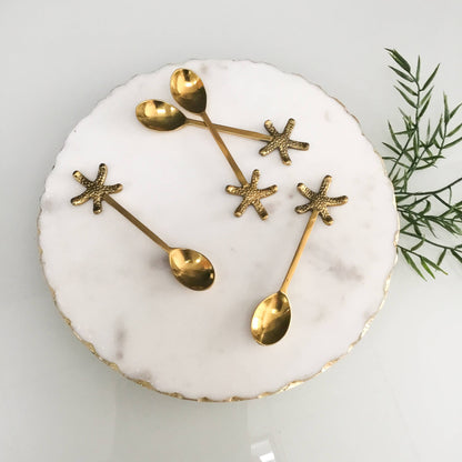 Set of gold spoons shaped like starfish on a white plate with a green leafy branch in the background.