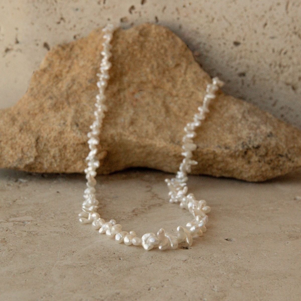 A delicate pearl strand necklace made of irregular white Keshi pearls draped over a large tan rock on a textured beige stone surface.