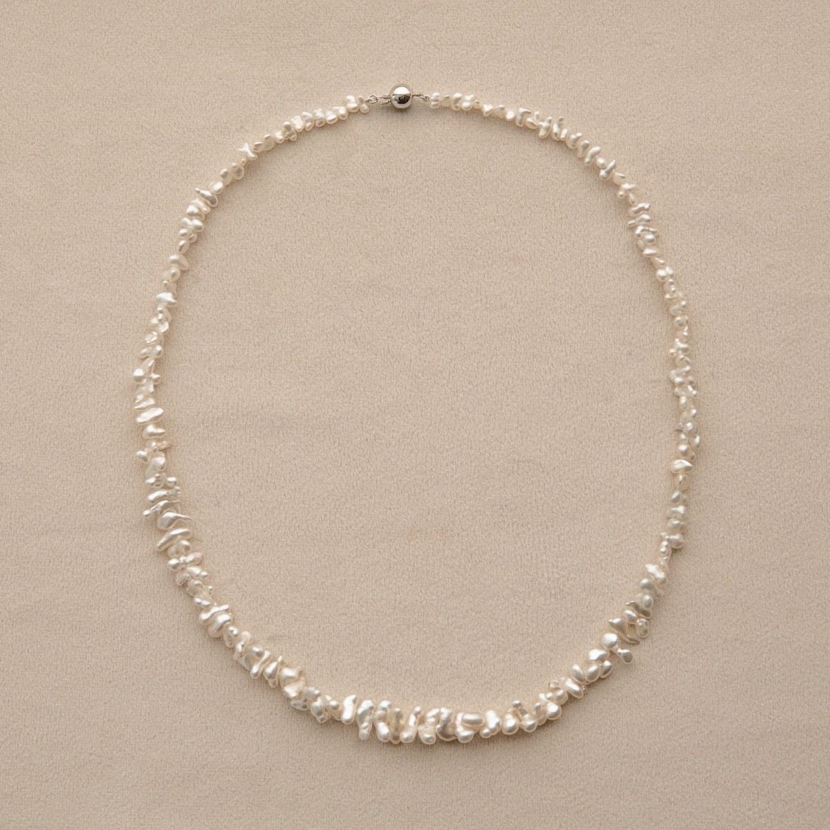 A delicate pearl strand necklace made of irregular white Keshi pearls arranged in a circle, displayed on a neutral beige background.
