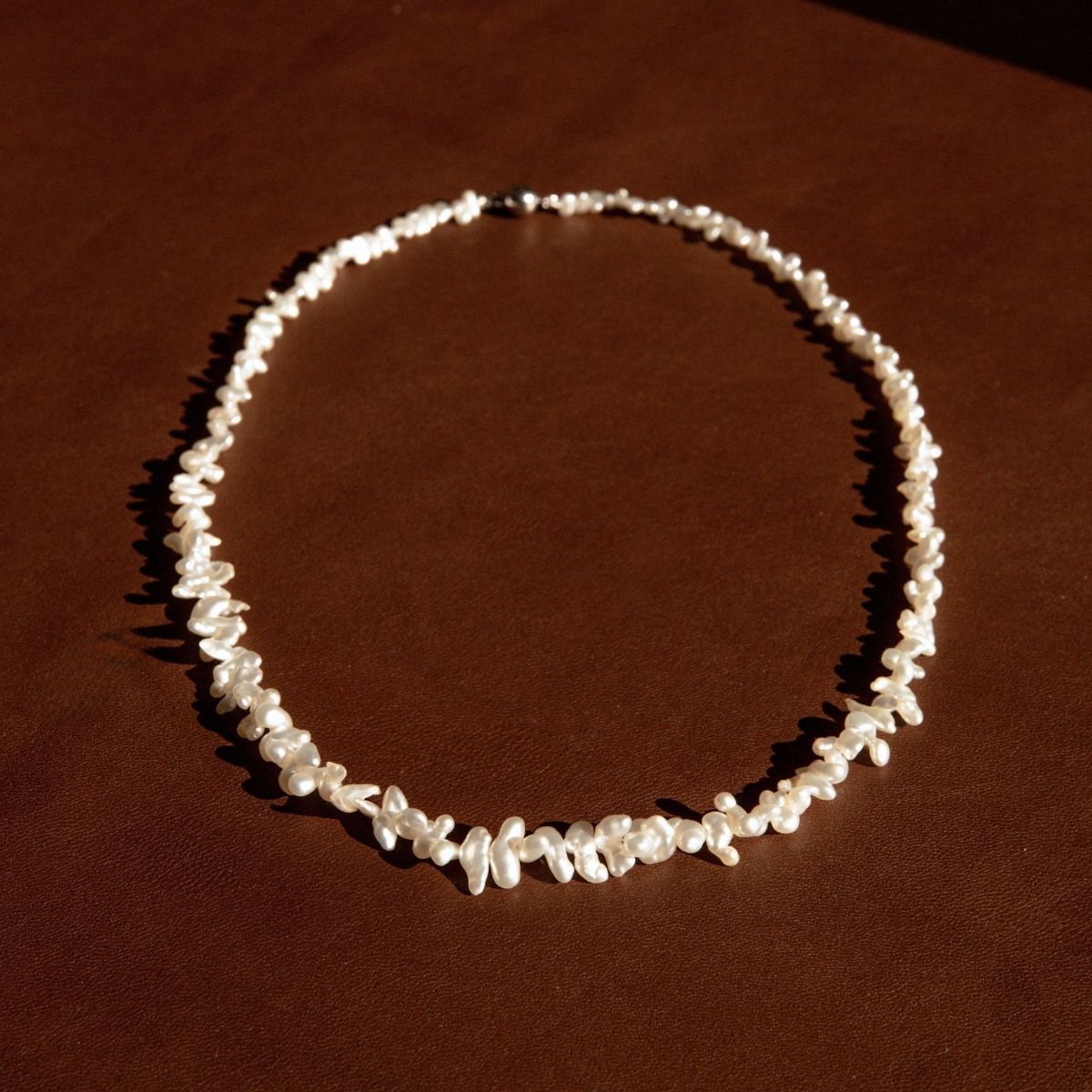 A necklace made of irregular Keshi pearls arranged in a circle, displayed on a dark brown textured surface under warm directional lighting.