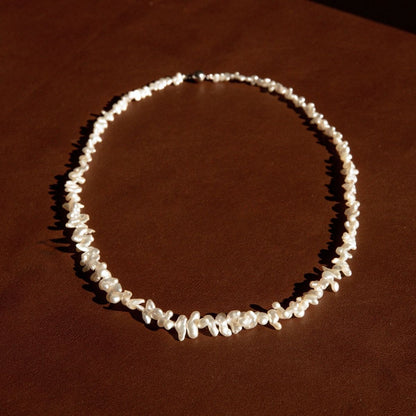 A necklace made of irregular Keshi pearls arranged in a circle, displayed on a dark brown textured surface under warm directional lighting.