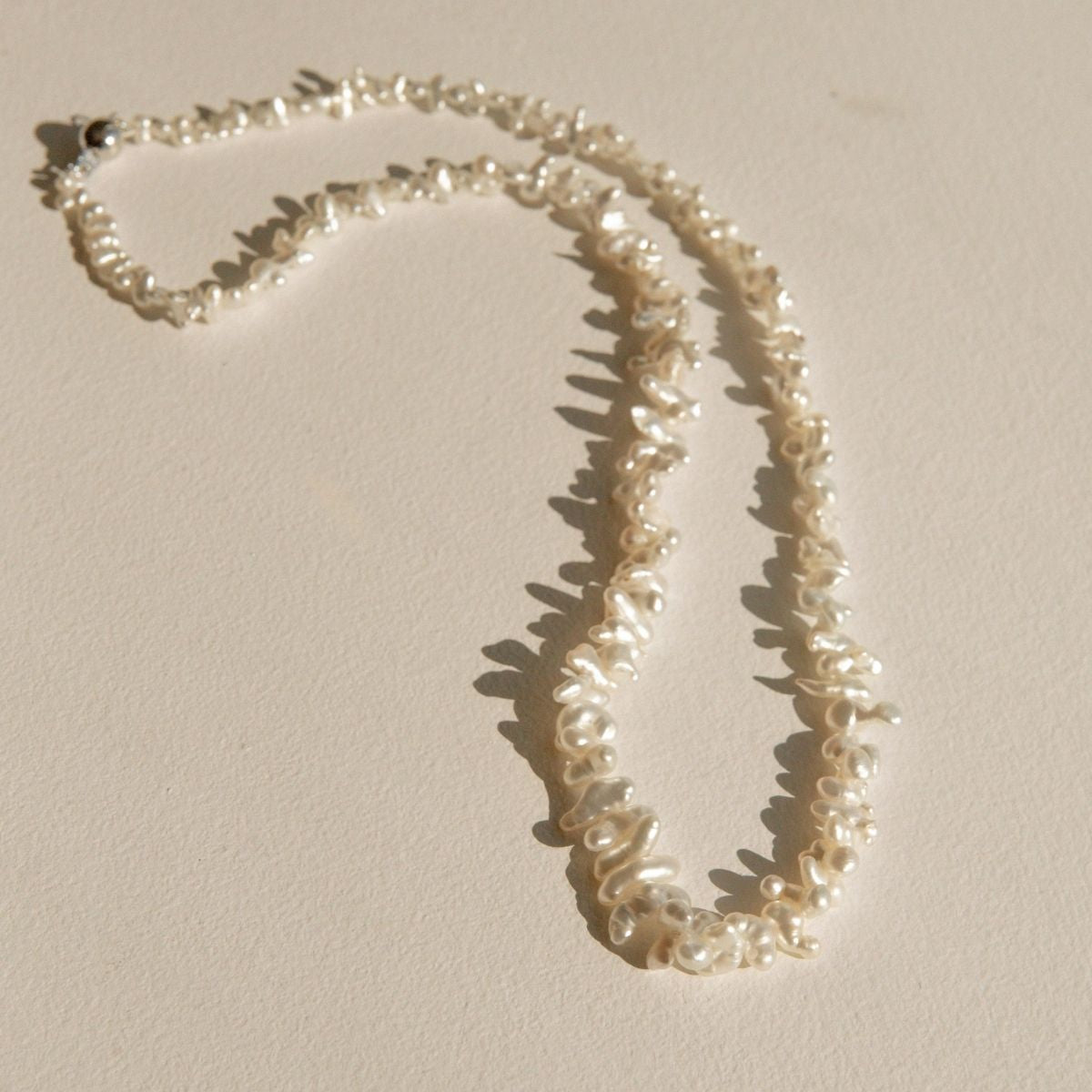 A strand necklace made of irregular, white Keshi pearls laid out on a warm beige surface with soft shadows.