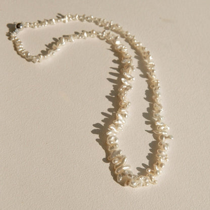A strand necklace made of irregular, white Keshi pearls laid out on a warm beige surface with soft shadows.