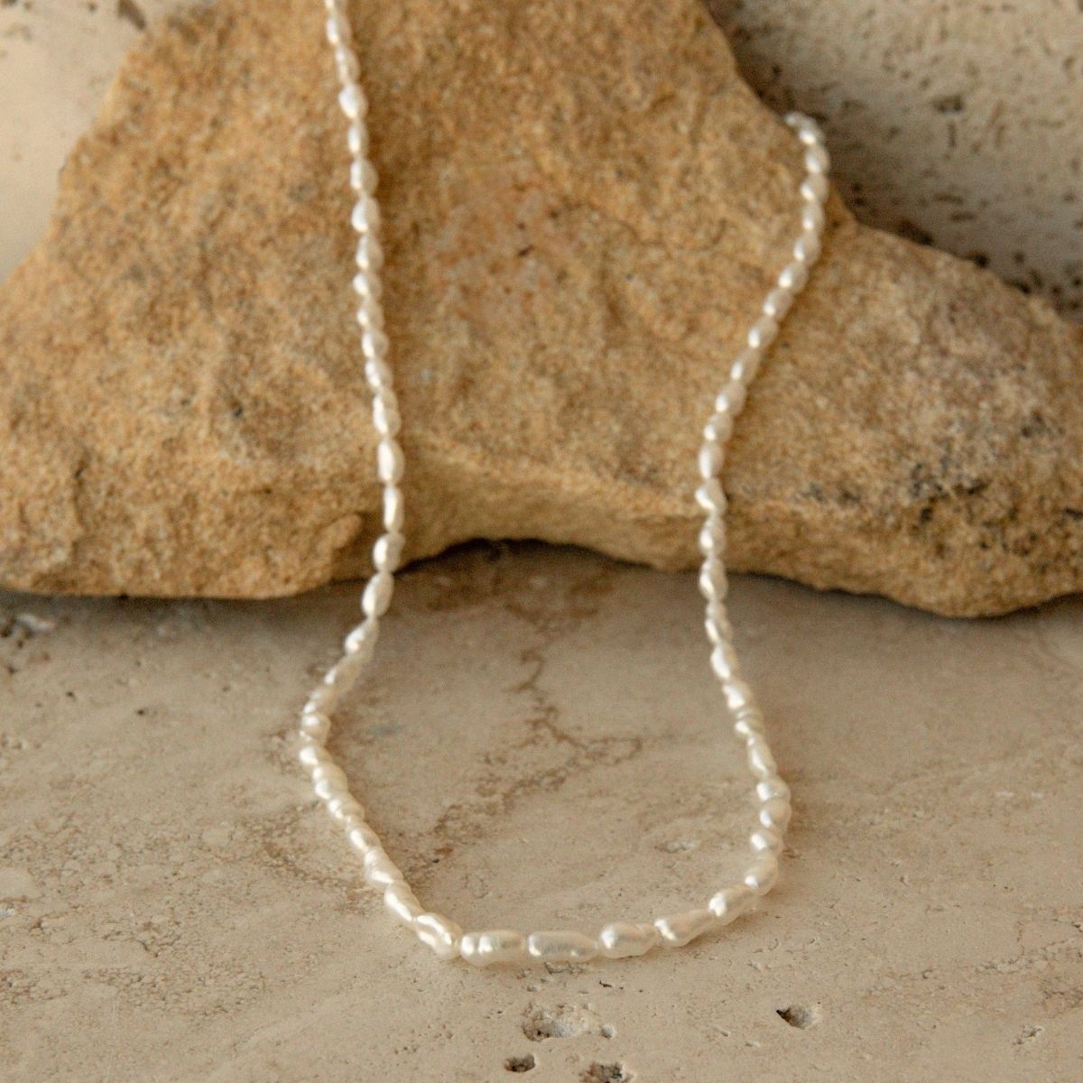 A delicate white Keshi pearl strand necklace draped over a natural tan rock on a textured stone surface.