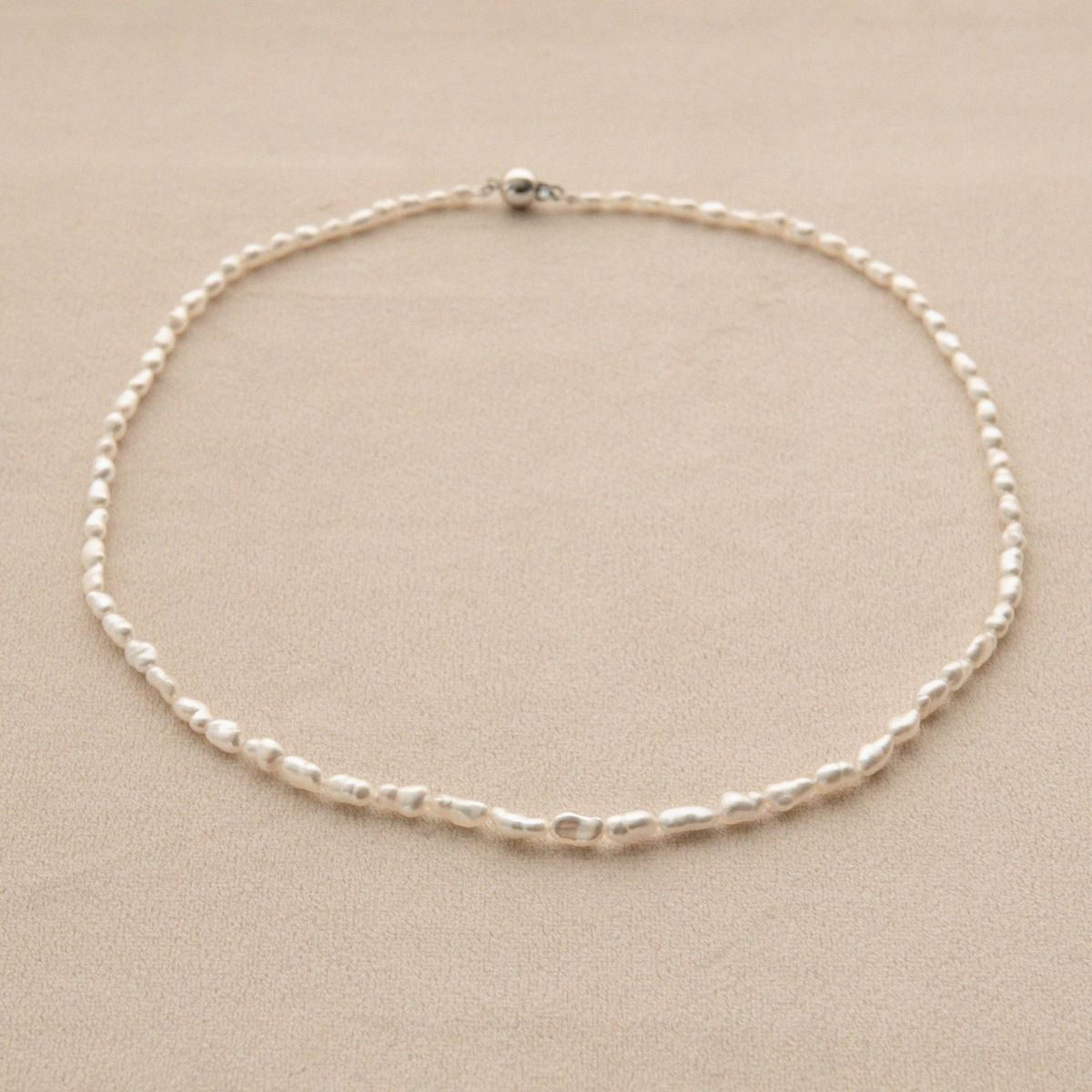 A delicate pearl strand necklace with irregular, organic shaped Keshi pearls arranged in a circle on a neutral beige background.