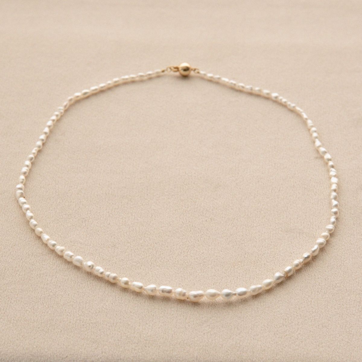 A delicate Keshi pearl strand necklace with a yellow gold clasp, displayed on a textured beige surface.