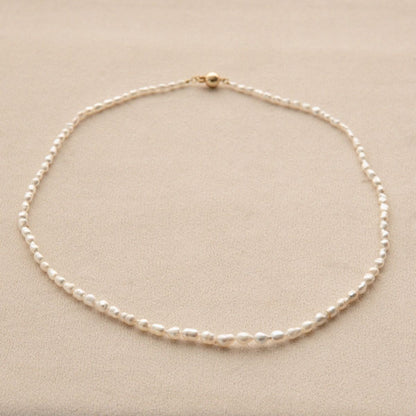 A delicate Keshi pearl strand necklace with a yellow gold clasp, displayed on a textured beige surface.