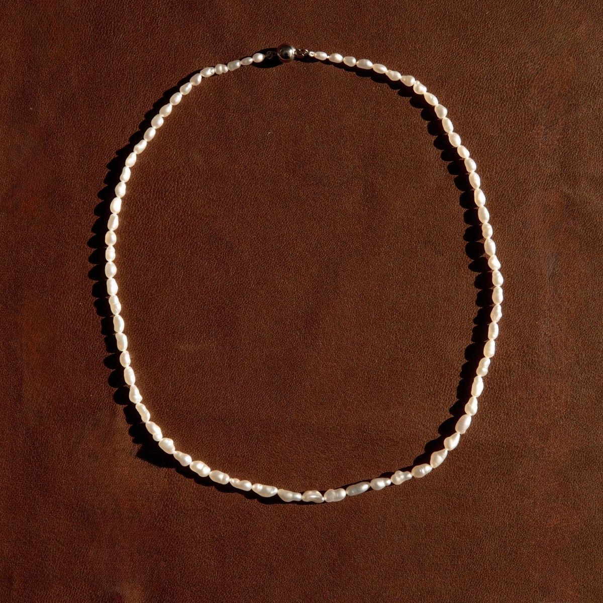 A single-strand Keshi pearl necklace arranged in an oval shape on a textured brown leather background.