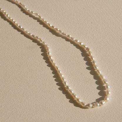 A delicate pearl strand necklace made of small irregular cream-colored Keshi pearls, arranged in a loose loop on a textured beige surface with soft shadows.
