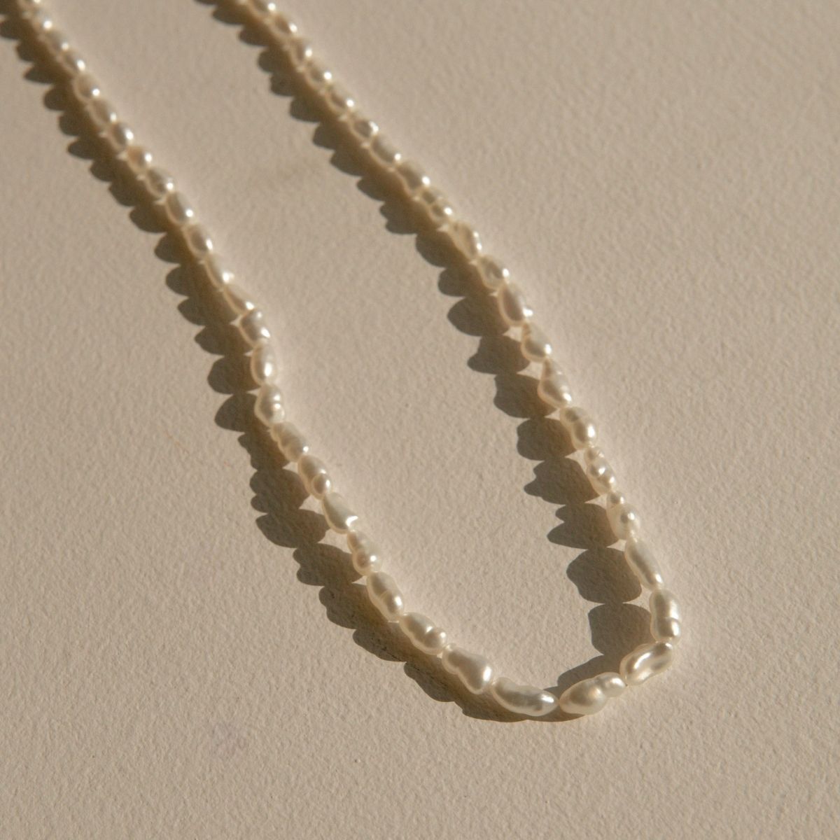 A strand of irregular white Keshi pearls draped in an U-shape on a textured beige surface, casting soft shadows.
