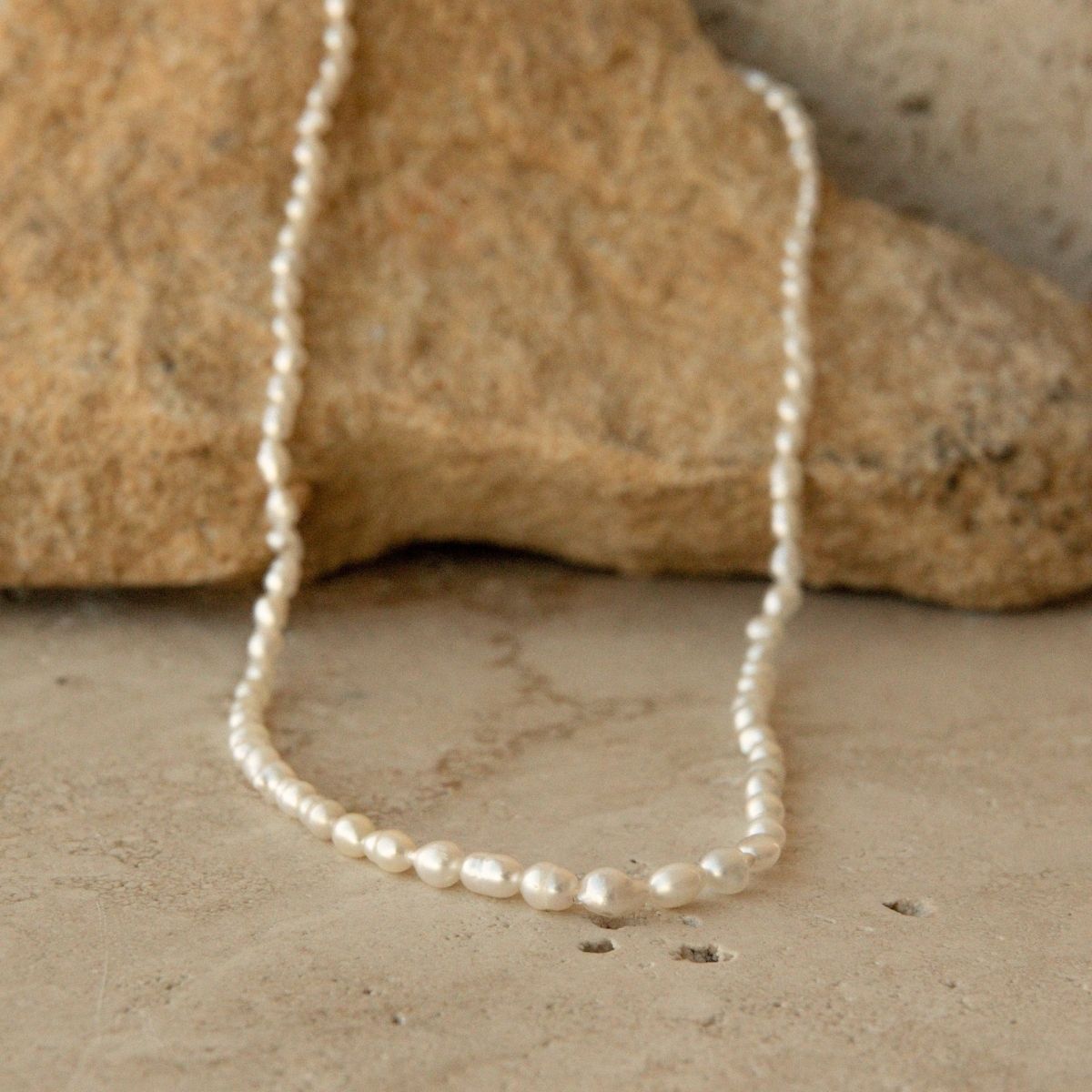 A strand of irregular white Keshi pearls draped over a tan stone on a textured beige surface.