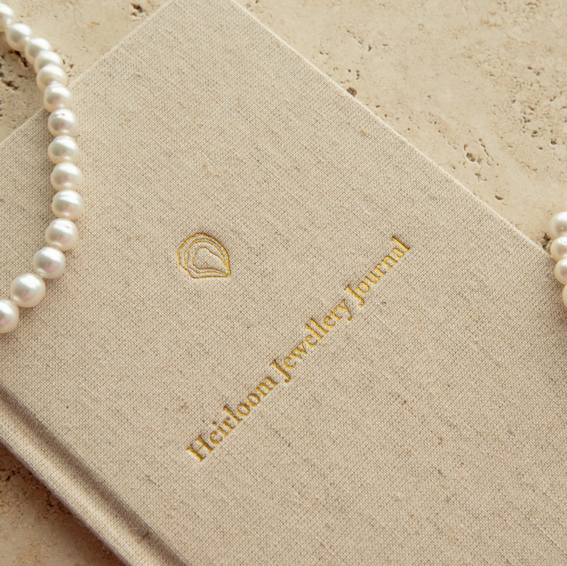 A close-up of a beige linen-covered journal with gold foil branding reading 'Heirloom Jewellery Journal,' styled with pearl necklaces and a pearl bracelet on a light stone surface.