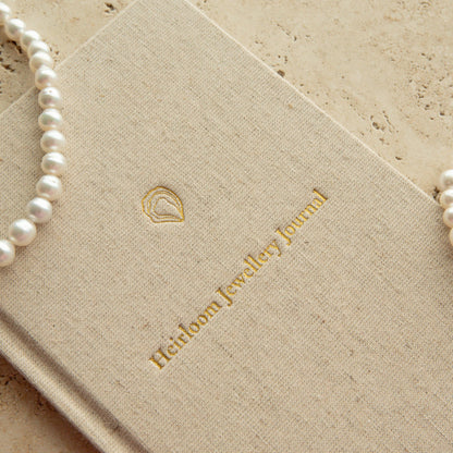 A close-up of a beige linen-covered journal with gold foil branding reading 'Heirloom Jewellery Journal,' styled with pearl necklaces and a pearl bracelet on a light stone surface.