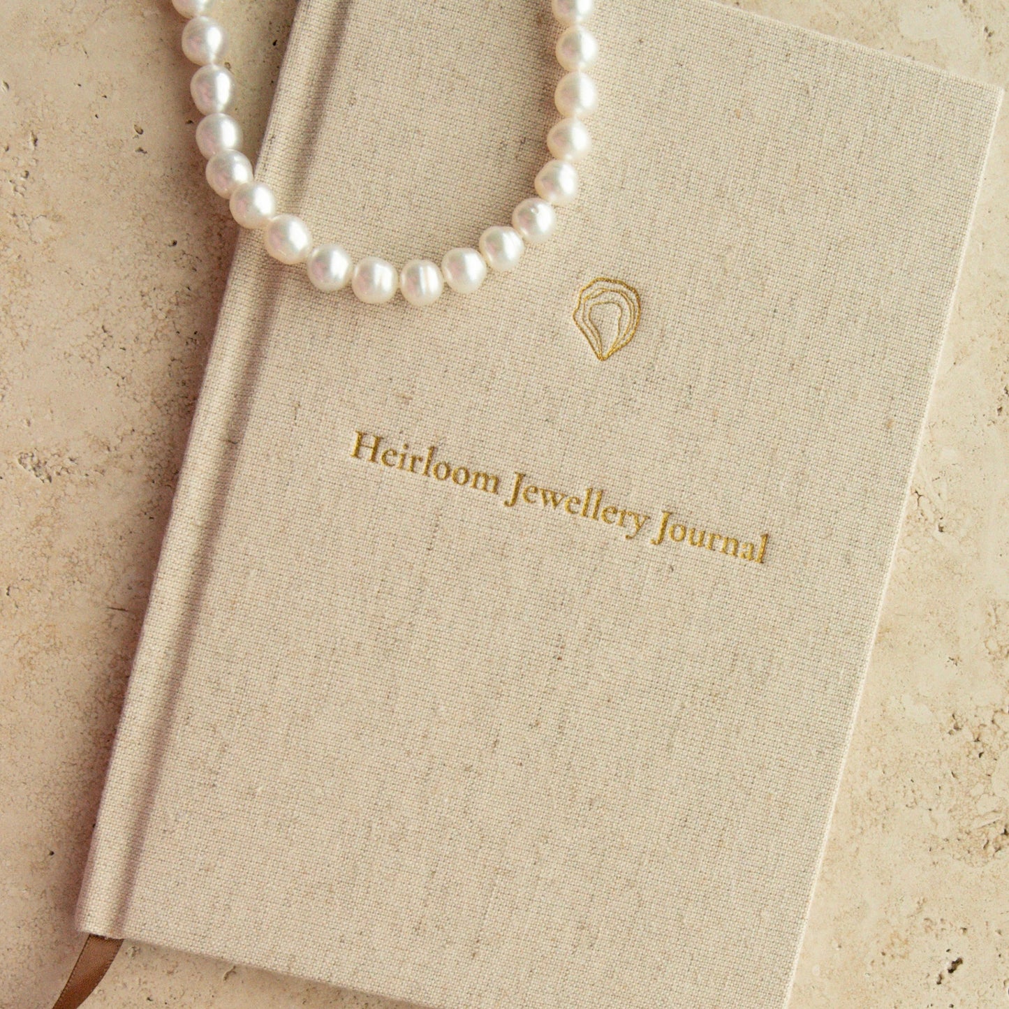 A cream-colored textured hardcover journal with gold foil lettering reading 'Heirloom Jewellery Journal', styled with white pearl strand necklace and a pearl bracelet on a beige stone surface.