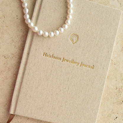 A cream-colored textured hardcover journal with gold foil lettering reading 'Heirloom Jewellery Journal', styled with white pearl strand necklace and a pearl bracelet on a beige stone surface.