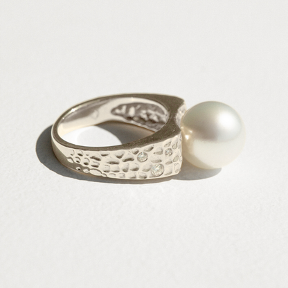 Silver ring with textured design and a pearl on a light gray background