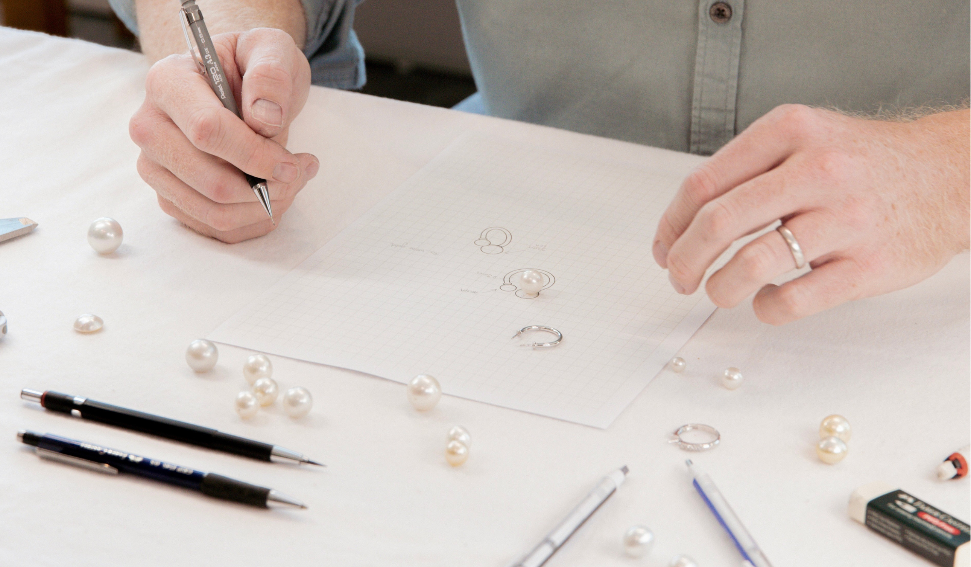 Person arranging pearls on a table, drawing timeless jewellery designs with pens and an eraser.