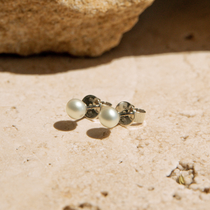 Pair of keshi pearl stud earrings on a sandy surface with a rock in the background