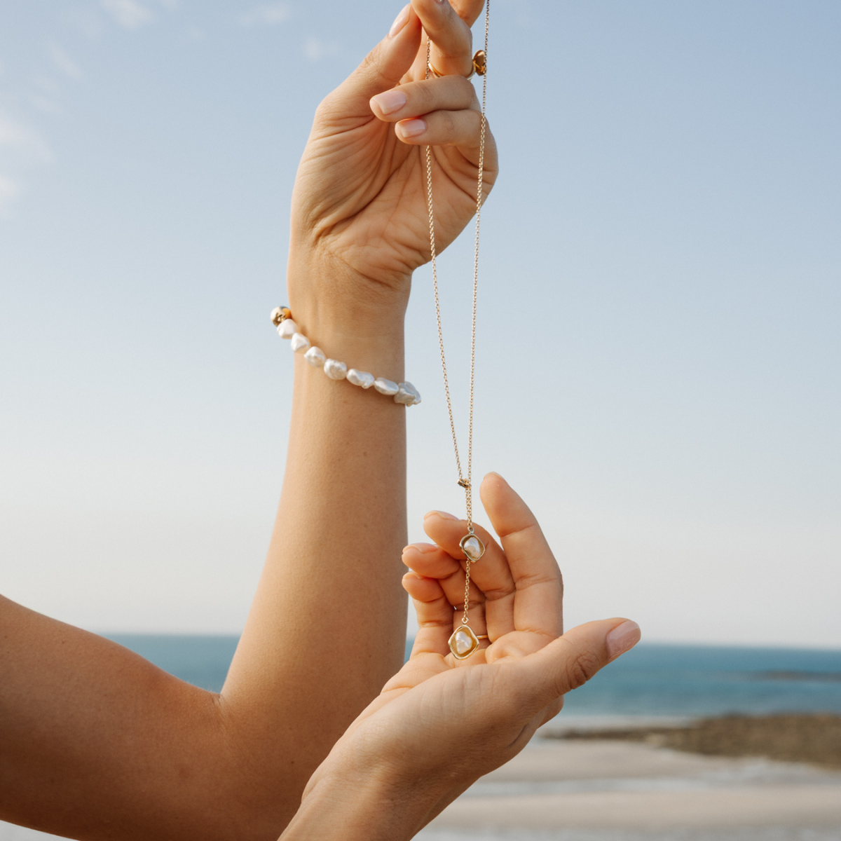 Hand holding a necklace with a pearl pendant against a beach background