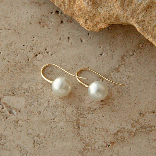 A pair of gold hook earrings with round white Australian South Sea pearls, displayed on a textured beige stone surface with a rock in the upper corner.