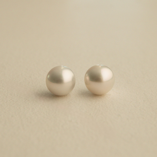 A pair of round, lustrous Australian South Sea pearls in a warm ivory or champagne tone, resting on a textured beige surface.