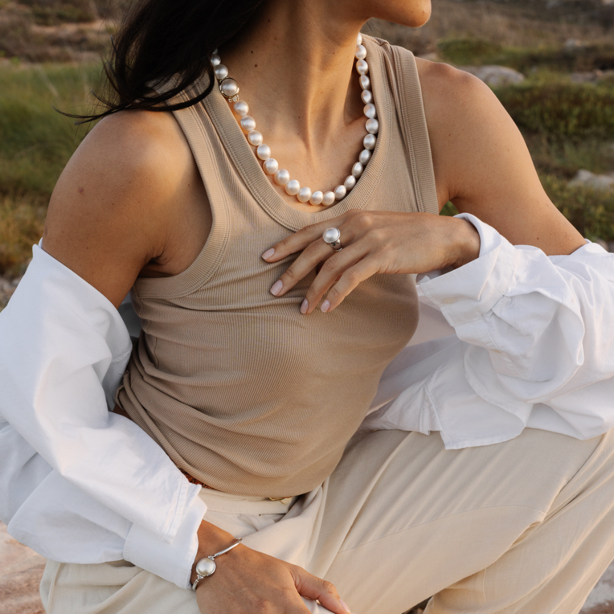 Woman wearing a beige top with a white blouse underneath, sitting outdoors. She wears a Mabe pearl strand necklace with matching mabe pearl jewellery pieces.