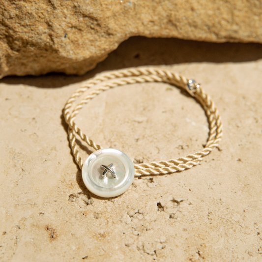 Bracelet with a Mother of Pearl button and sand coloured silk cord on a sandy surface