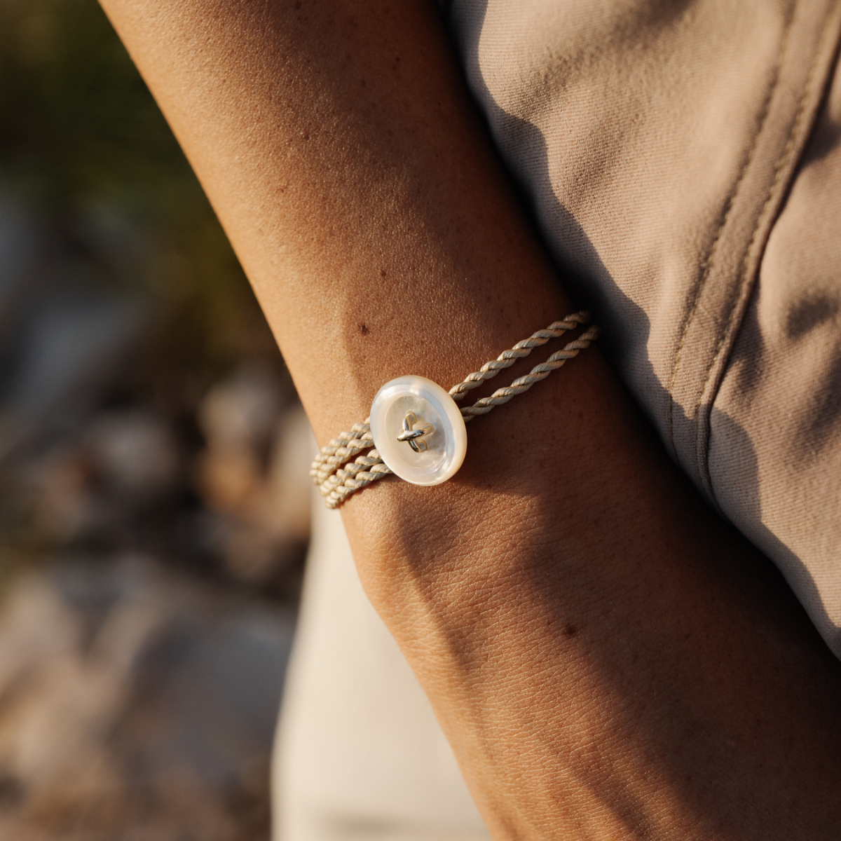 Close-up of a wrist wearing a silk bracelet with a Mother of Pearl button charm, against a blurred natural background.