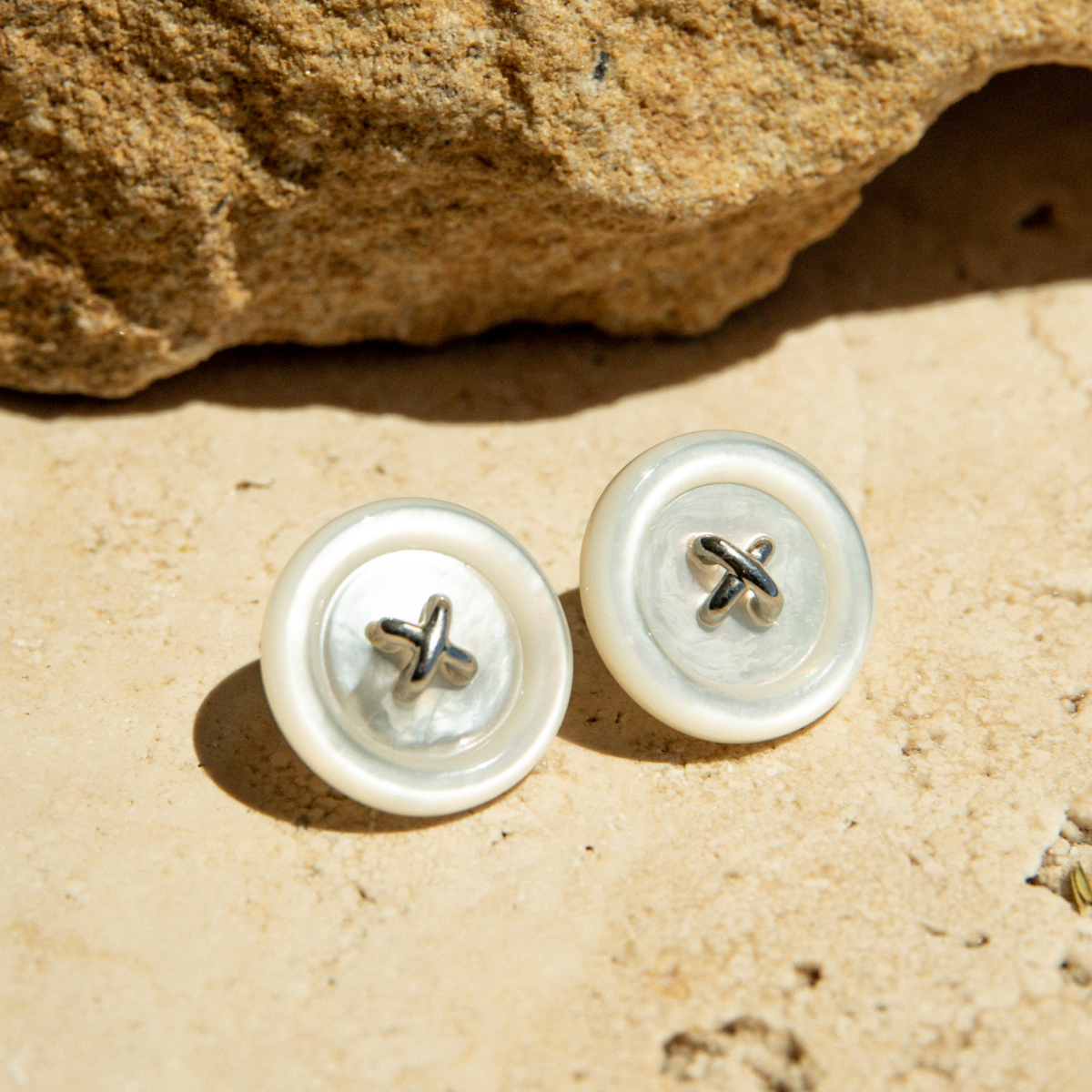 Pair of Mother of Pearl earrings on a sandy surface with a rock in the background