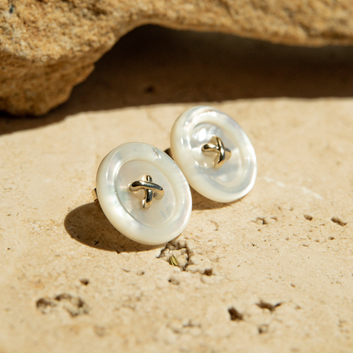 Mother of Pearl stud earrings on a sandy surface with rocks in the background