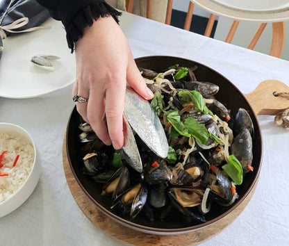 Person grabbing mussels with their Mussel Shovel in a bowl on a table with a white background