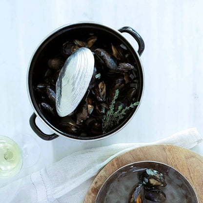 Pot of mussels and mussel shovel with a serving on a wooden board, glass of white wine on the side.