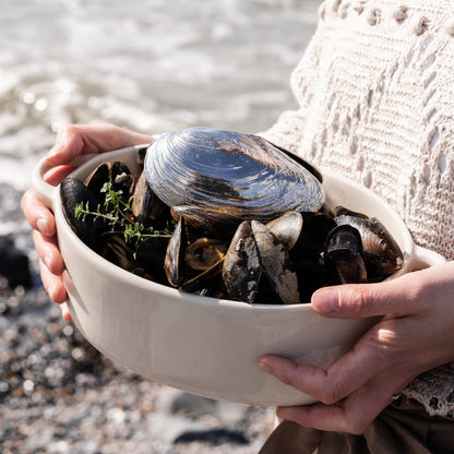 Person holding a bowl filled with mussels and mussels shovel by a body of water.