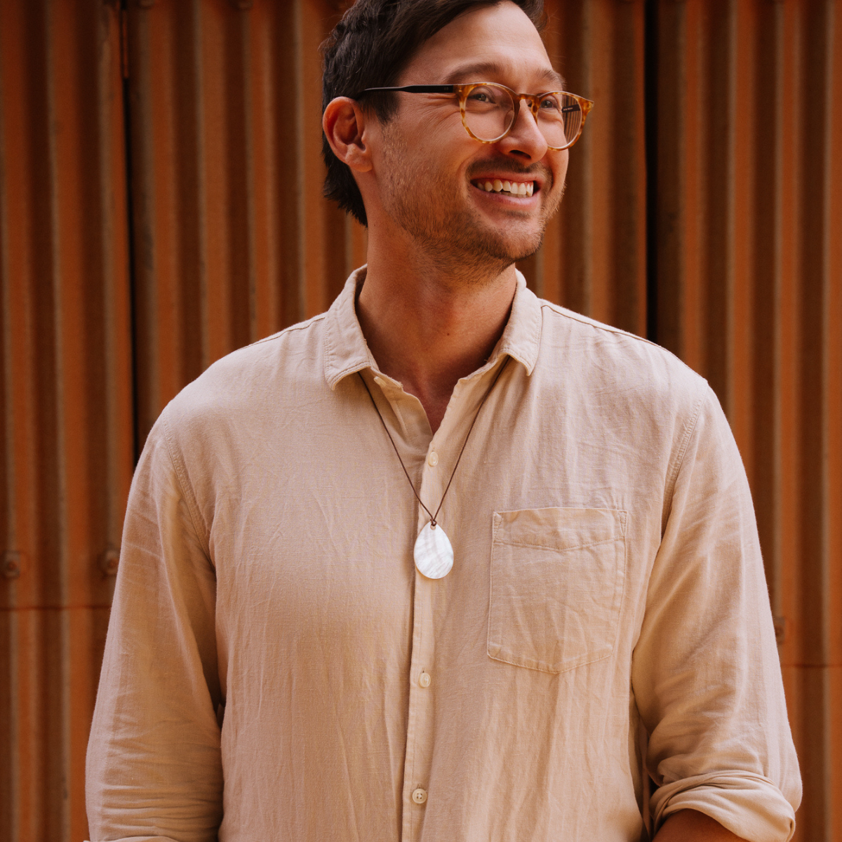 Man wearing glasses and a beige shirt with a mother of pearl pendant, standing against a wooden paneled wall.