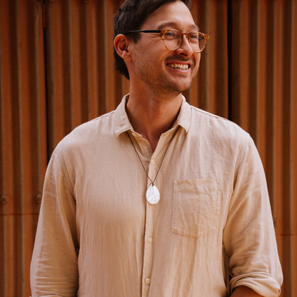 Man wearing glasses and a beige shirt with a mother of pearl pendant, standing against a wooden paneled wall.