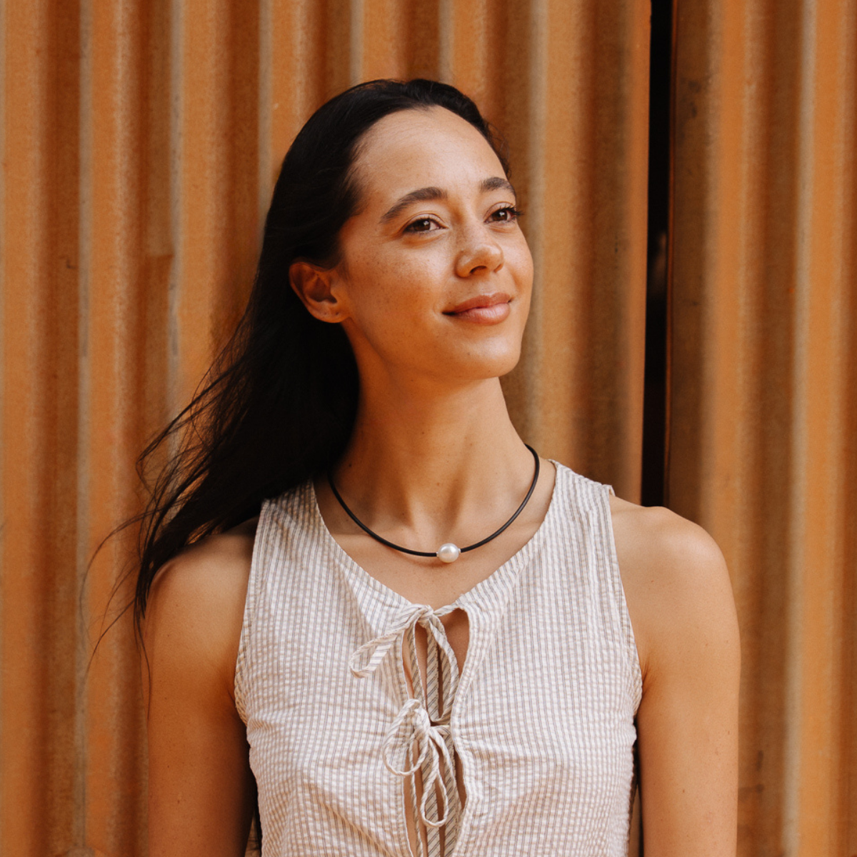 Woman with long dark hair wearing a sleeveless top and neoprene pearl necklace against a wooden background