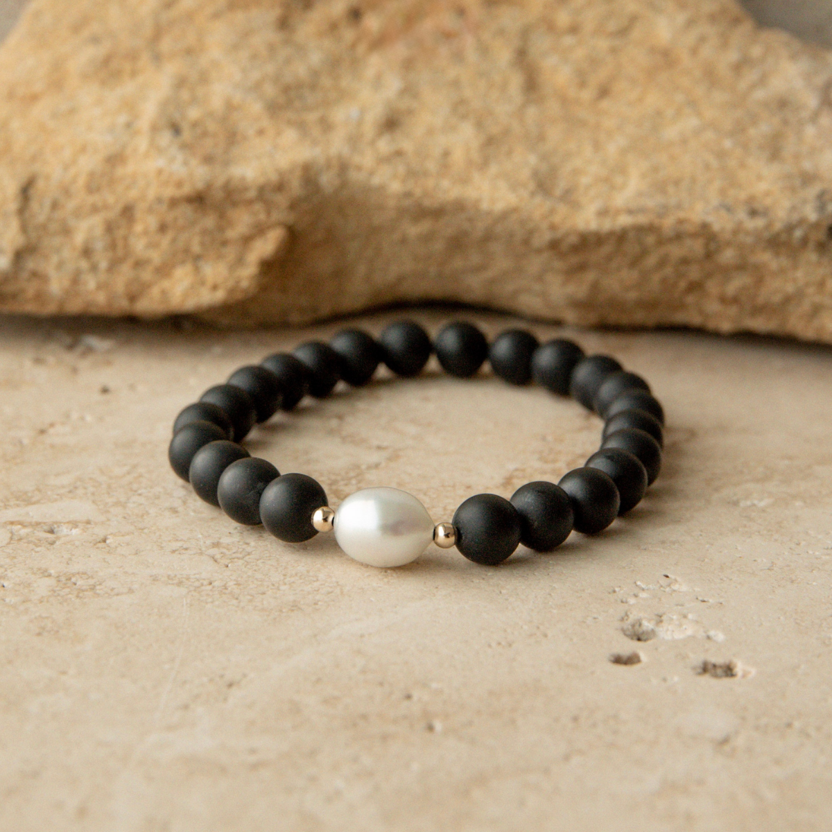 Black beaded bracelet with a white pearl on a beige surface with a rock in the background