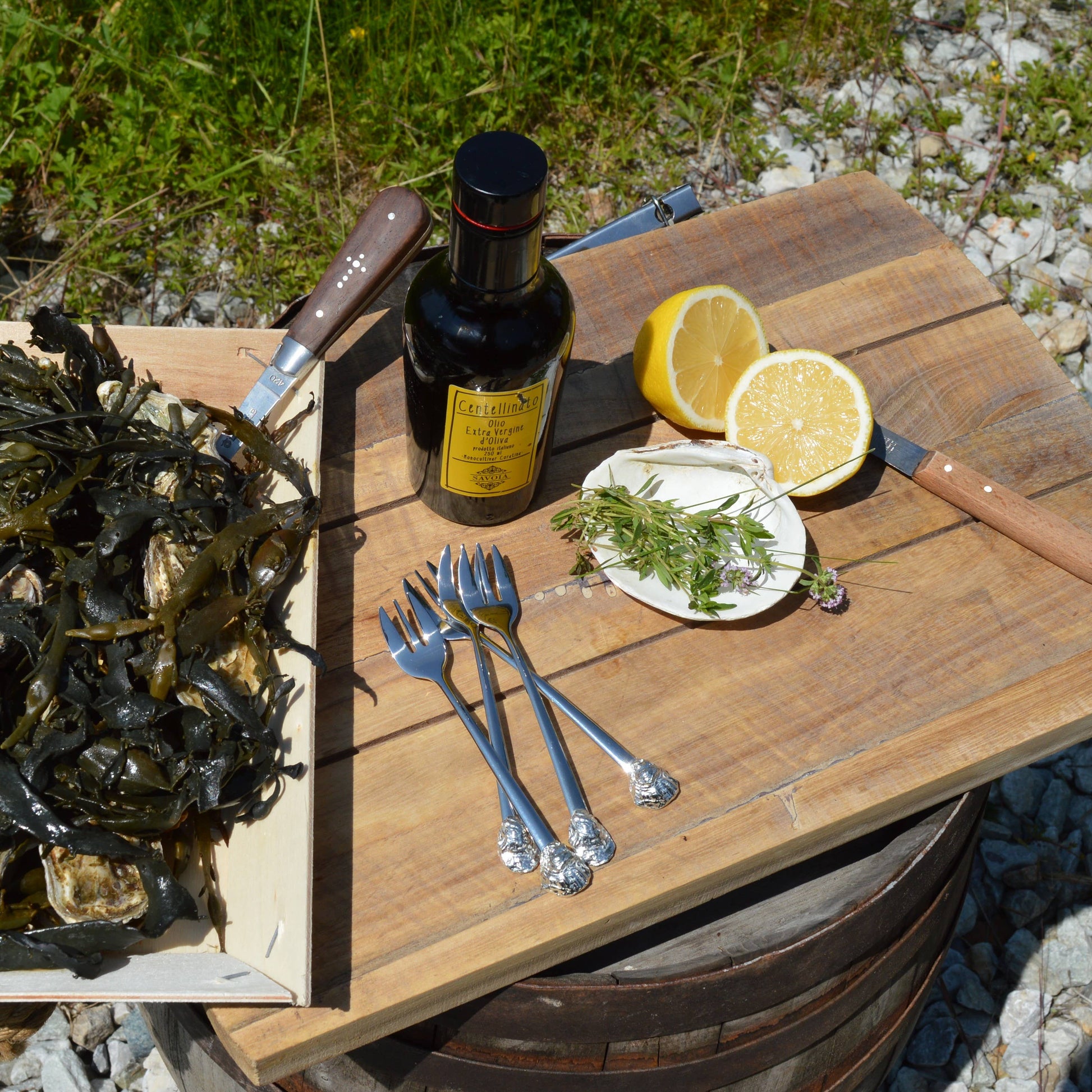 Bottle of olive oil, lemons, and utensils on a wooden board outdoors.
