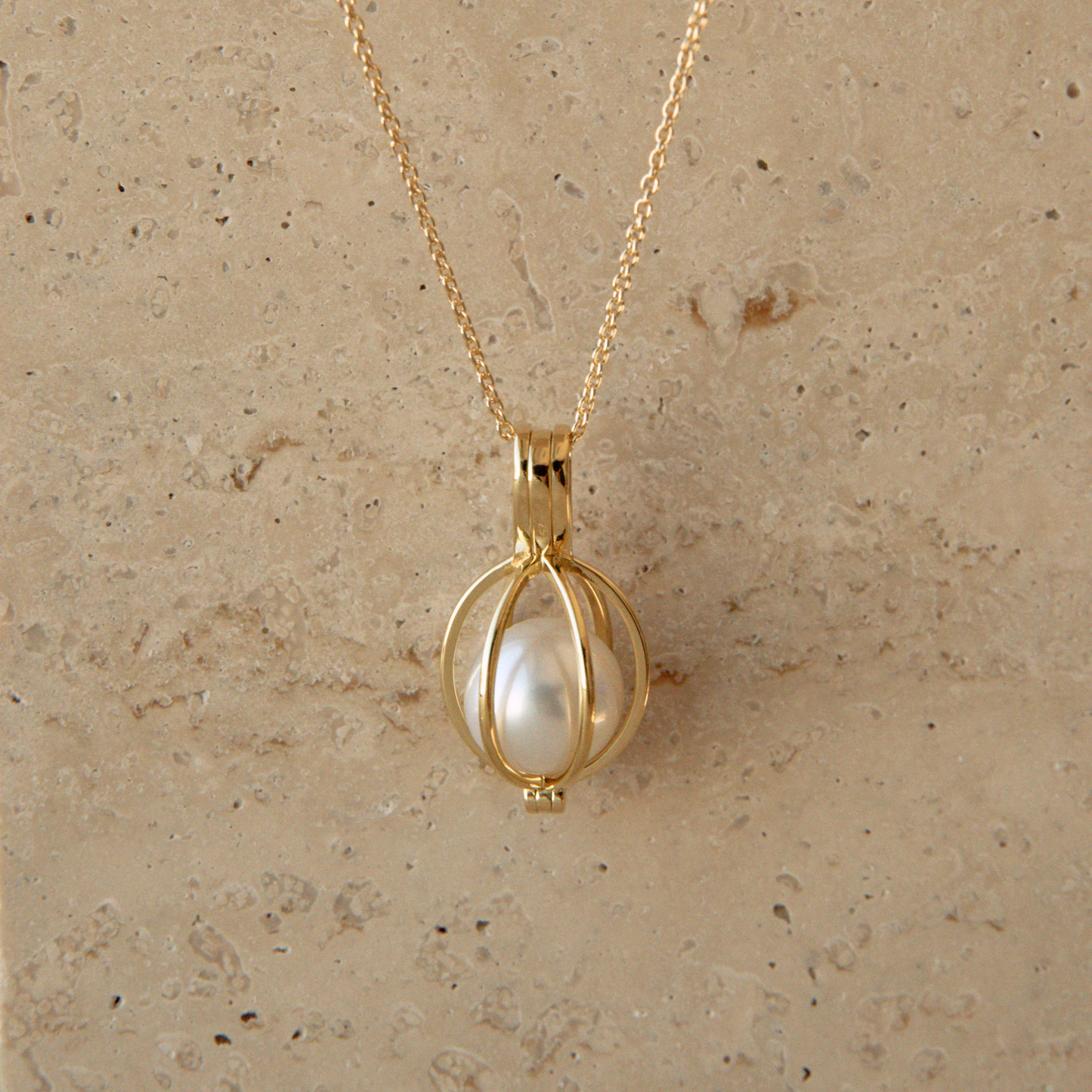 A gold chain necklace with a spherical gold cage pendant holding a single white Australian South Sea pearl, displayed against a textured beige stone background.