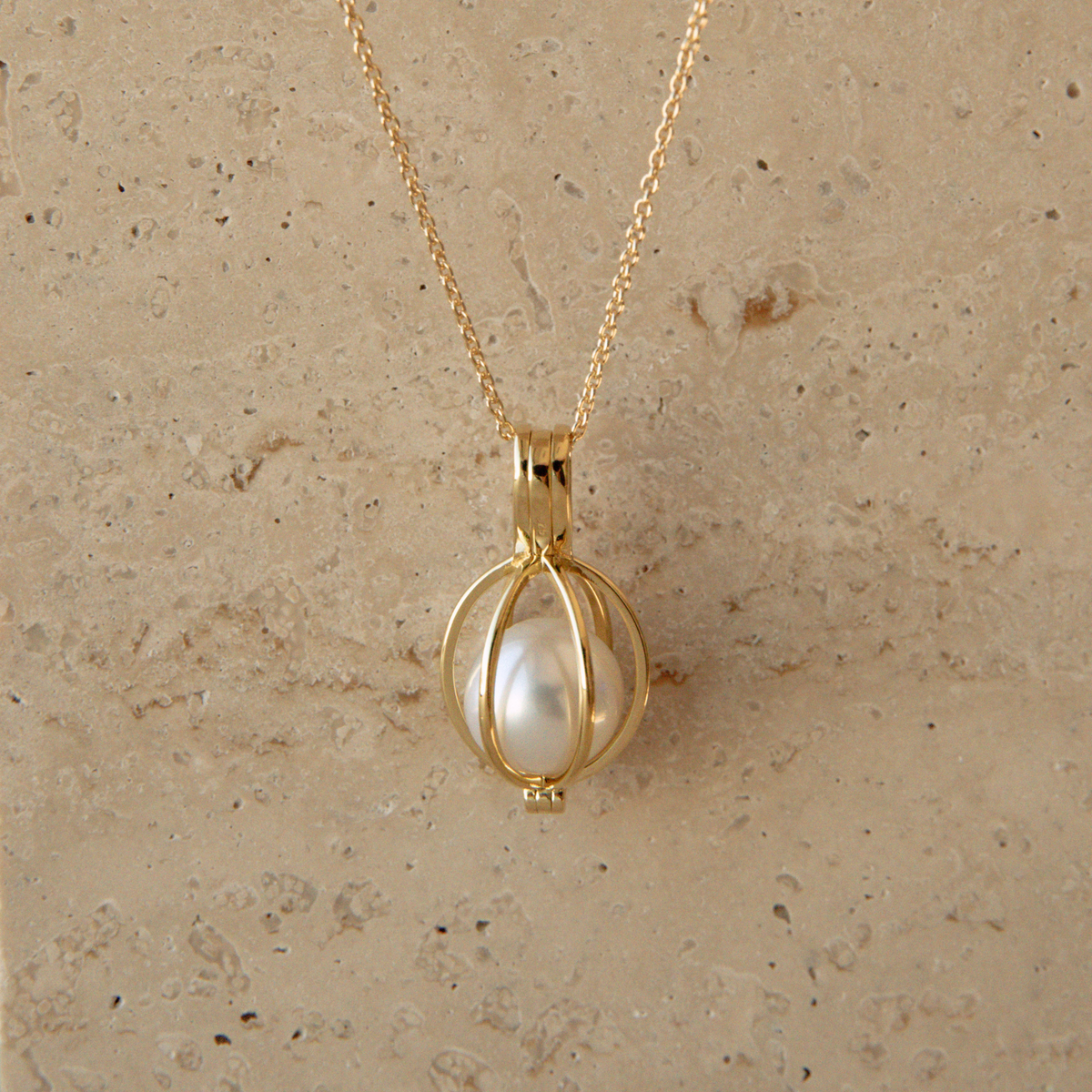 A gold chain necklace with a spherical gold cage pendant holding a single white Australian South Sea pearl, displayed against a textured beige stone background.