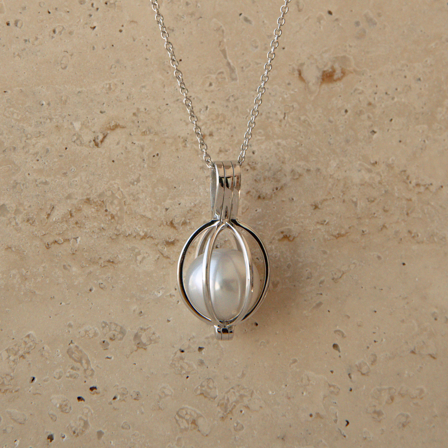 A silver pendant necklace featuring a white Australian South Sea pearl enclosed in a circular cage-like setting, hanging from a delicate silver chain against a textured beige stone background.