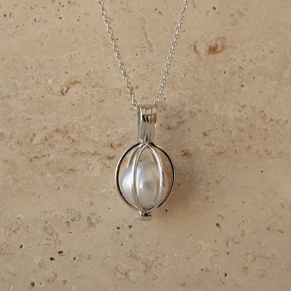 A silver pendant necklace featuring a white Australian South Sea pearl enclosed in a circular cage-like setting, hanging from a delicate silver chain against a textured beige stone background.