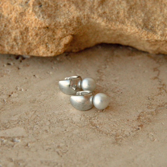 A pair of silver-toned stud earrings with small round Australian Akoya pearls, photographed on a sandy surface with a rock in the background.