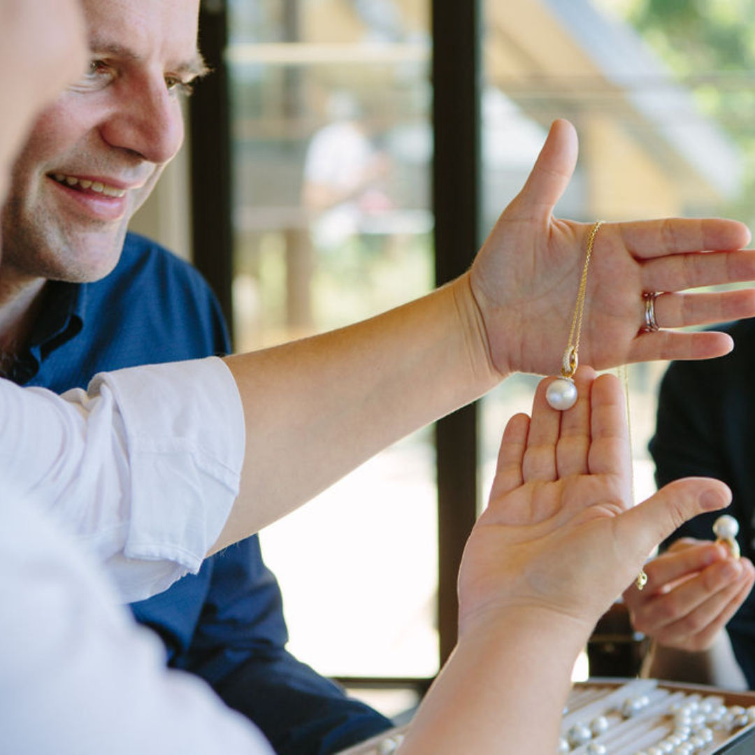 A couple inspect a beautiful Australian South Sea pearl jewellery piece, set in gold and complemented with white sparkling diamonds during their visit at our Shellar Door in Broken Bay. They both smile at the spectacular beauty of this gorgeous pearl jewellery piece.