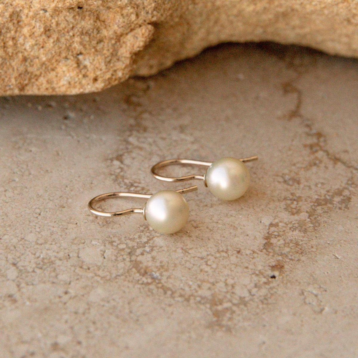 A pair of gold hook earrings with round white Australian Akoya pearls, displayed on a textured beige stone surface with a rock in the background.