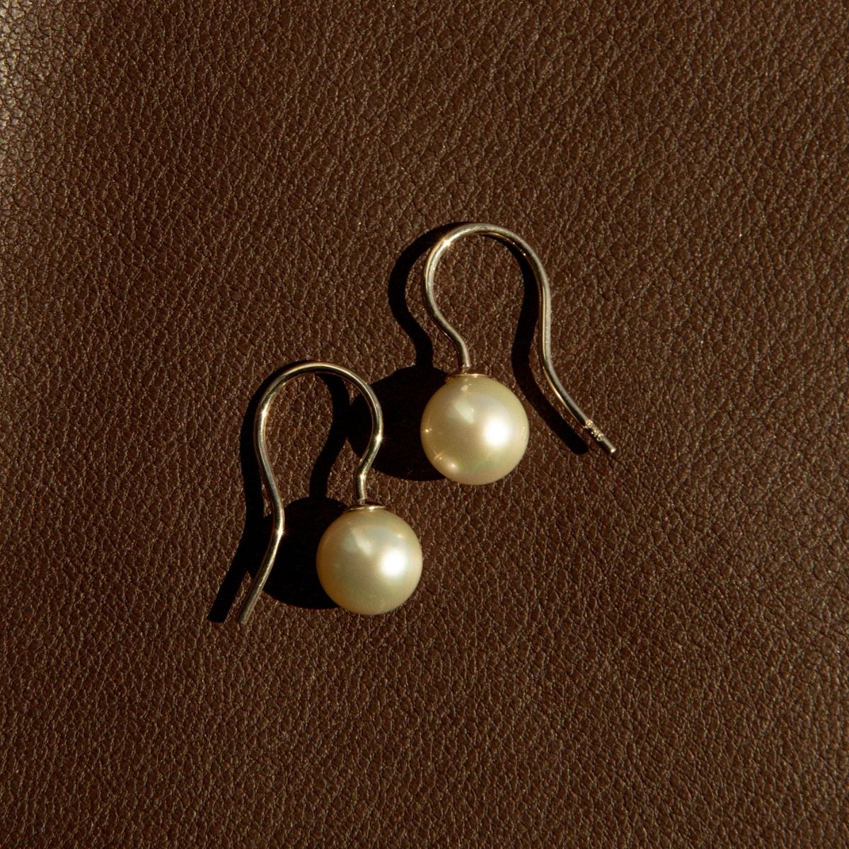 A pair of hook earrings with round cream-colored Akoya pearls and silver-toned hooks, displayed on a textured brown leather surface.