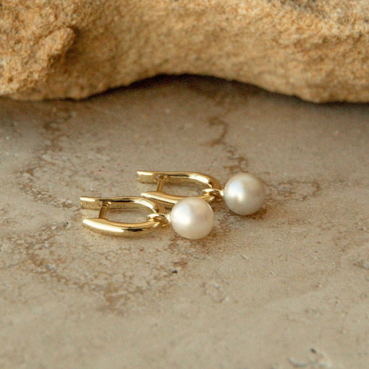 A pair of gold huggie earrings with round white Australian Akoya pearls, displayed on a textured beige stone surface with a rock in the background.