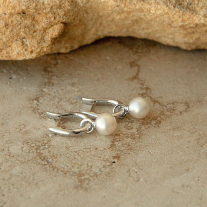 A pair of silver huggie earrings with small round white Australian Akoya pearls, displayed on a textured beige stone surface with a rock in the background.